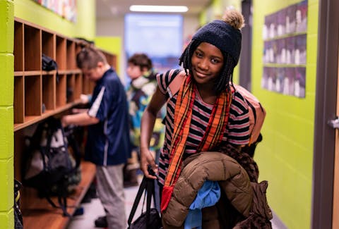 Smiling middle schooler standing in the school hallway while holding a jacket and other personal items